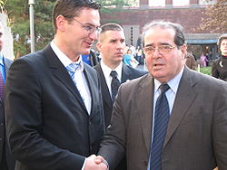 Скалия, одетый в бежевый пиджак поверх рубашки и галстука, пожимает руку Юрию Топлаку из Европейской ассоциации избирательного права, ожидая взгляда в камеру.