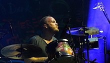 A bald, bearded man wearing a black shirt plays the drums