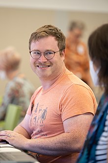 A man with glasses and an orange t-shirt is sitting and smiling at someone off-camera