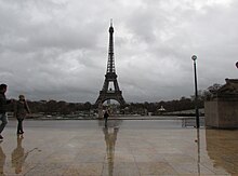 Esplanade of the Trocadero in Paris after rain. The layer of water exhibits specular reflection, reflecting an image of the Eiffel Tower and other objects. Eiffel Tower under cloudy sky.jpg