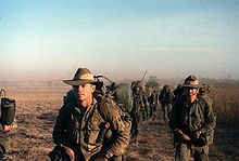 Um grupo de homens vestindo uniformes militares verdes andando por terreno árido.
