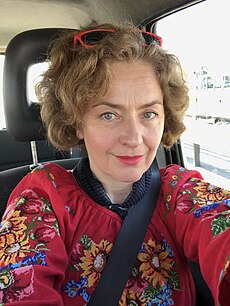 Laryssa Artjuhina ist auf einem Selfie im Auto sitzend zu sehen. Ihr rötliches halblanges gelocktes Haar wird von einer roten Sonnenbrille gehalten. Sie trägt ein rotes Oberteil mit bunten Blumenmotiven und schaut direkt in die Kamera.