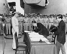 Un homme en tenue de soirée se penche sur une table pour écrire sur un papier.  Un homme blanc en uniforme le regarde.  Un Japonais en vêtements formels regarde par derrière.  En arrière-plan, de nombreux hommes blancs en uniforme observent.