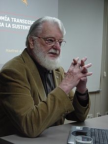 Manfred Max-Neef se sienta en una mesa cerca de una computadora portátil.  En la pared detrás de él hay una diapositiva de una presentación.