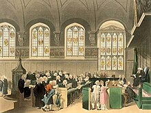 A courtroom seen from the side. Made of stone, there is a large, arched ceiling and four stained glass windows. There is a single wigged judge, assisted by a bench of clerks in front of him. In front of the clerks sit two rows of barristers, in black gowns and curled white wigs.