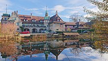 L'immagine mostra la fattoria Riedern, una villa che è stata costruita nello stile di un maniero del XIX secolo e il riflesso dell'edificio in un prato antistante.