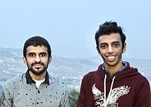 Zwei junge Männer, einer im Hemd und einer in einem burgunderfarbenen Hoodie, vor der Kamera