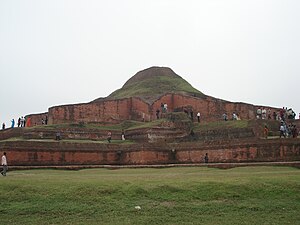 Ruins of a structure of red stone now resembling a small hill or mound.