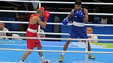 Deux boxeurs olympiques, l'un en bleu, l'autre en rouge, combattent face à face sur un ring surélevé.