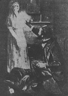 image noir et blanc : deux personnes à gauche une jeune femme en blanc, à droite un homme accroupi levant le bras vers la jeune femme