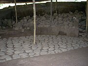 An area of excavation under a corrugated roof supported by wooden poles. The foreground shows an even cobbled pavement running diagonally from left front to right back, where it disappears into a vertical wall of partially excavated rubble and soil.