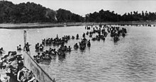 Britse soldaten waden aan wal vanaf landingsvaartuigen.  Op de achtergrond is een met bomen omzoomd strand