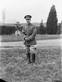 A man in a neat military uniform with a peaked cap, ribbons on his tunic, Sam Brown belt, cane, riding boots and spurs.