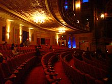 curved rows of red seats, decorative ceiling gold under the circle, pillars, a few people seated waiting