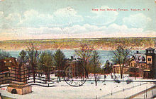 A postcard showing a view over a body of water in the distance in winter, with snow on the ground. There are bare trees and some houses in the foreground