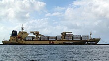 Photographie du flanc tribord d'un navire cargo, à bonne distance. Il est jaune et chargé de containers blancs qui portent le logo de la marque.