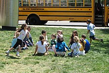 Un grupo de niños está sentado en círculo sobre el césped y dos más corren alrededor del círculo uno tras otro.