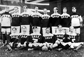 Photographe d'une équipe de football posant devant un grand drapeau canadien dont on devine le Red Ensign. Les joueurs portent un maillot bicolore estampillé GALT