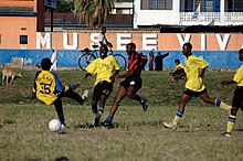 cinco homens jogando futebol, uma camisa listrada vermelha e preta e quatro em camisa amarela