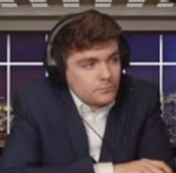 Nick Fuentes, pictured from a webcam. He is wearing a white shirt and navy sport coat, and the background is a photograph of a cityscape