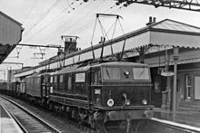 Black-and-white photo of electric locomotive with overhead wiring at a station
