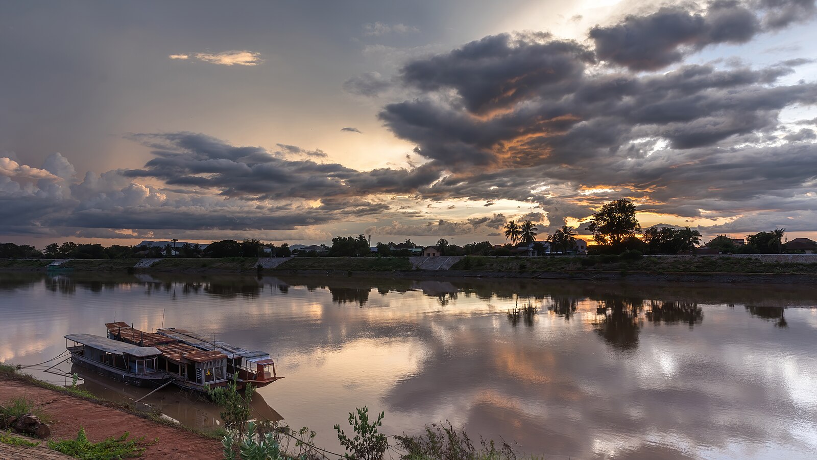 Water reflection of sunset with gray and orange clouds and boats moored to the bank in Pakse Laos