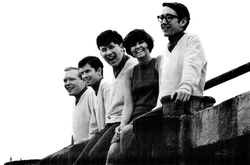 Four young men wearing light-coloured sweaters and a young woman in a dark-coloured dress, all sitting on a wall