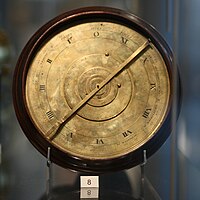 Photo of a clock-like mechanical device in a museum display case, with a small card with the number 8 printed on it