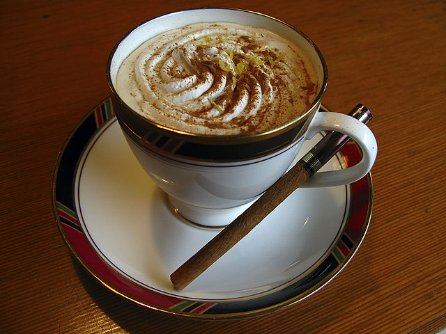 כוס קפוצ'ינו עם ציור בקפה עליו ומבחש הקינמון לצידו.