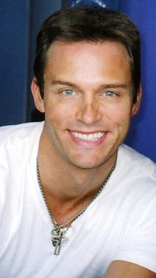 A man with dark hair wearing a white T-shirt including a silver necklace.