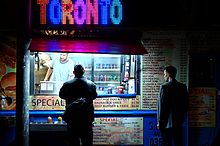 A man stands in front of a fast food cart in Toronto at night. The sign on the cart says Toronto in blue, pink, and purple neon lighting.