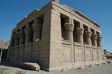 Small stone building surrounded by half-walls and columns with elaborate capitals