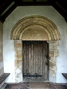 Простая деревянная дверь, над которой находится круглая арка с резным тимпаном.