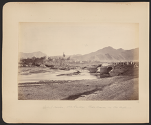 One of five bridges that crossed Kabul River during the Second Anglo-Afghan War (1879-1880) era.  Soldiers a pictured atop the bridge while people walk along the road in the distance and in the right foreground people sit or squat on the bridge while soldiers ride behind them. Bala Hissar (High Fort) is in the background just visible through the heat haze and trees. It was the locus of power in Kabul for many centuries and the site of fierce fighting during the war. It was partly destroyed in October–December 1879 when Sir Frederick Roberts occupied the city at the head of the Kabul Field Force