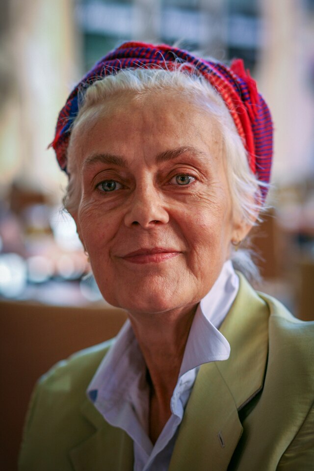 Une femme aux cheveux blancs, aux yeux clairs, portant un foulard dans les cheveux, regarde l'objectif avec un demi-sourire.