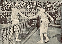 Lenglen and Wills shaking hands at the net while an official at the side holds their right hand up in a "stop" gesture