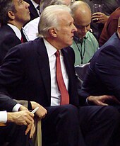Um homem de cabelos brancos, vestindo um terno preto, camisa branca e gravata laranja, sentado em um jogo de basquete.