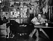 Man sitting cross-legged at table, pipe in hand, apparently writing. Much clutter of clothing, books and equipment is in the background.