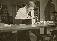 Black and white photograph of Zita Stead, in a white lab coat, seated at a desk and looking down a microscope.