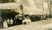Cliché noir et blanc. Des convives sont assis sous une tente blanche, à une table dont on ne voit pas les extrémités.