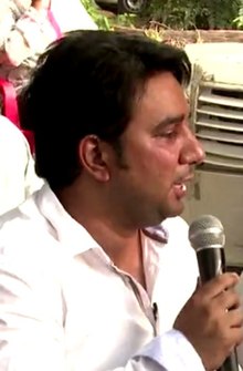 South Asian male, wearing white shirt and holding microphone, looking left of camera