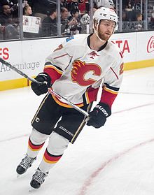 A hockey player with a red beard skates on an ice rink. He is wearing a white jersey with a stylized red C on the front and is holding a hockey stick in both hands.