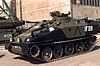 A small tracked armoured personnel carrier with black and green camouflage pattern, parked on a paved surface. Other tanks and a building are in the background.