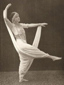 A young white woman in a white costume with an exposed midriff and bare feet, posed with both arms and one leg outstretched, and her foot pointed