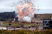 Viewed from a distance, with a telephoto lens, a large explosion is captured in its early stages. In the foreground, assorted building materials are visible. In the background, a hillside is partially covered by a forest.