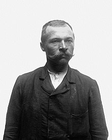 Photographie en noir et blanc d'un homme, en buste, portant un costume et de longues moustaches.