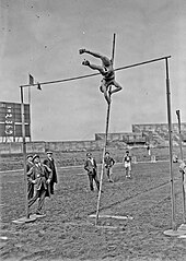 Photographie en noir et blanc d'un homme sautant à la perche dans un stade sous le regard d'hommes en costumes.