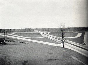 "A black and white photograph of a divided roadway being crossed by another roadway. The horizon cuts the photo in half. The setting is mostly rural, with the roadways slicing through that. The divided road extends from the lower right corner to the centre of the horizon, while the second roadway crosses horizontally halfway towards the foreground. Connecting the two separated roadways are a series of ramps. Although only half visible from the angle of the photo, the ramps form the shape of a four-leaf clover surrounded by a diamond."