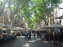 Pedestrians walk between trees and cafe seating (18793606571).jpg