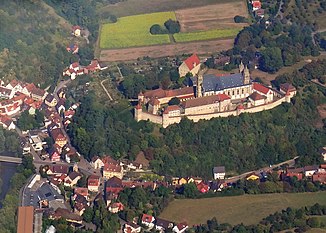 Blick auf das unterste Waschbachtal in Steinbach und die Comburg auf ihrem Umlaufberg gegenüber. Der Bach fließt entlang der Straße in der Talrinne von rechts nach links, teilweise verdolt, ab der Dorfkirche bis zur Mündung an der Brücke am linken Bildrand wieder offen.